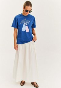 Modrá tričko s krátkymi rolovanými rukávmi, s grafikou ružových papúč a textom. V kombinácii s bielou, tečúcou maxi sukňou a obuvou s leopardím vzorom.