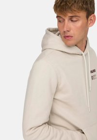 Sudadera beige con cordón, bolsillo frontal y pequeño detalle de logo impreso; hecha de un tejido suave y texturizado, con puños acanalados.