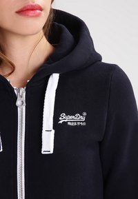 En ung person som bär en marinblå SuperDry hoodie med vita dragsnören och en framzipper, läppar synliga med en naturlig rosa färg.