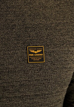 Dunkelgrüner strukturierter Strickstoff mit einem gelben und schwarzen Etikett, auf dem „PME LEGEND“ und „GETESTET IN HOHER HÖHE“ steht.