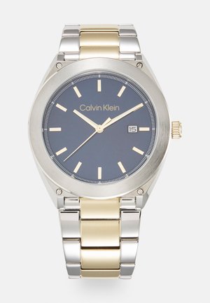Montre en acier inoxydable argent et or avec un cadran gris rond, aiguilles dorées et une fenêtre de date à 3 heures. Nom de la marque affiché.