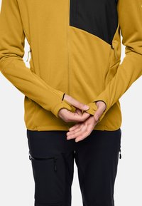 Giacca in pile gialla con pannello nero sul petto, dotata di tasche con zip e fori per il pollice sui polsini. Indossata sopra pantaloni neri con tasche.