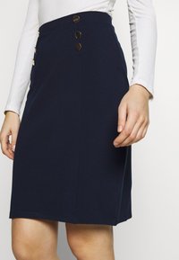 Námořnická modrá sukně do kol s přizpůsobeným střihem, na boku zdobená třemi zlatými knoflíky a vyrobená z hladké, texturované tkaniny.