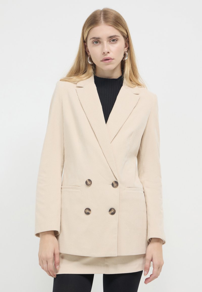 Blazer doppiopetto in corduroy beige con revers a punta, due tasche frontali e bottoni scuri. Indossato sopra un dolcevita a coste nera.