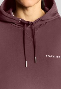 Bordeaux hættebluse med snoretræk, ribbet detaljering og sølvhardware-accents. Har "LYLE & SCOTT" logo i hvid. Glat tekstur.
