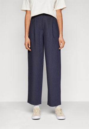 Personne portant un pantalon large à fines rayures bleu marine, des chaussettes blanches, des baskets beiges et un haut blanc à manches courtes, debout devant un fond uni.