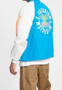 Bundička s barevným blokováním, modré tělo, bílé rukávy a grafický potisk na zadní straně. Vyrobena z lehkého materiálu s volným střihem.