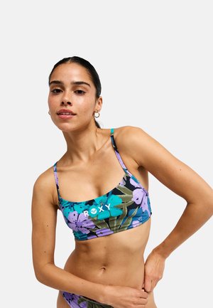 Jonge vrouw stelt paarse en turquoise bloemenbikinitop met dunne bandjes af tegen een effen lichte achtergrond.