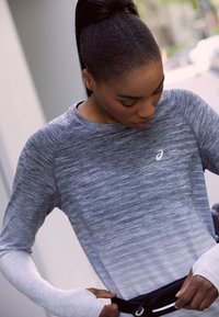 Grå atletisk langærmet skjorte med et struktureret mønster, der har hvide ærmer som accent. Skjorten har et lille logo nær halsudskæringen og en taljeelastik.