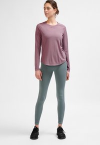 Langärmliges lila Sportoberteil mit geschwungener Saumlinie, kombiniert mit figurschmeichelnden grünen Leggings. Schwarze Sportschuhe runden das Outfit ab.