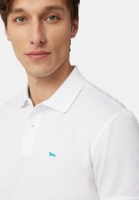 Polo bianco con un accento verde sul colletto e un piccolo logo ricamato turchese sul lato sinistro del petto. Maniche corte, vestibilità classica.