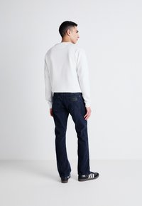 Sudadera blanca de cuello redondo, jeans azul marino y zapatos negros con rayas blancas. Enfoque en la vista trasera, mostrando un atuendo casual con un corte relajado.