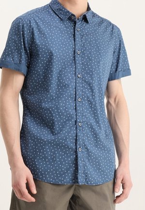 Homme portant une chemise bleue à manches courtes avec un petit motif blanc et un pantalon kaki sur un fond uni.