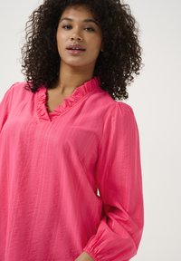 Blouse rose vif avec un col à volants et de longues manches bouffantes. Présente un tissu texturé avec de légères rayures verticales.