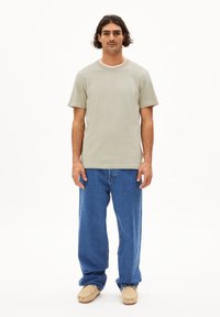 Hellgrünes Baumwoll-T-Shirt mit kurzen Ärmeln, kombiniert mit lockeren blauen Jeans und beigen Slippern. Schlichtes Design und neutrale Farben.