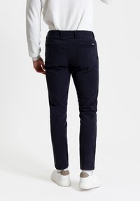 Chino blu navy con vestibilità slim, due tasche posteriori con zip e una texture liscia, indossati con sneakers bianche e un maglione leggero.