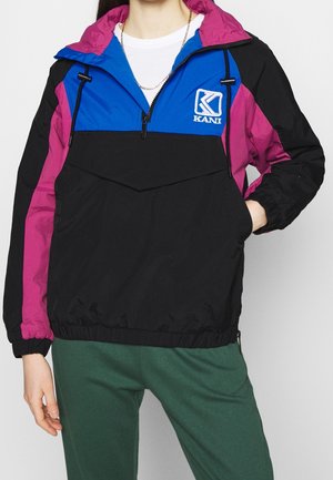 Personne portant une veste à blocs de couleurs avec des panneaux noirs, bleus et violets, fermeture éclair à l'avant et un logo Kani sur la poitrine, associée à un pantalon vert.