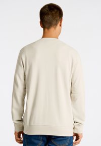 Sudadera de color beige claro con mangas largas, cuello redondo y puños acanalados. Textura suave, diseño sencillo, combinada con jeans azules.