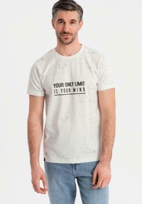 Camiseta blanca con un diseño texturizado, que presenta la frase "TU ÚNICO LÍMITE ERES TÚ MISMO" en negro, combinada con jeans azules.