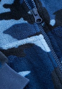Fleece jack met een blauw en zwart camouflagepatroon, voorzien van een marineblauwe ritssluiting en geribde manchetten, zachte textuur en variërende stofkleuren.