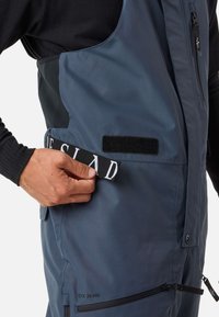 Blaue Außengarderobe mit matter Oberfläche verfügt über verstellbare Träger, eine mit Reißverschluss verschlossene Tasche und ein mit Klettverschluss befestigtes Logopatch.