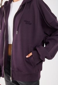 Sweatshirt roxa com fecho éclair, capuz ajustável com cordão, punhos canelados e bolso; apresenta logo da marca bordado no peito; textura de tecido suave.
