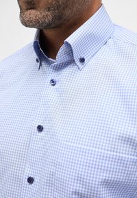 Camicia a righe azzurro chiaro e bianco con colletto aperto, dotata di bottoni blu scuro e tasca sul lato sinistro.