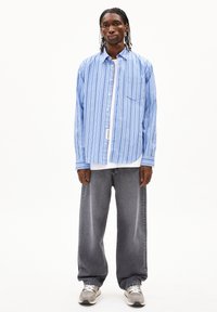 Blauwe verticaal gestreepte shirt met een zak en een knoopsluiting, gedragen over een wit shirt, gecombineerd met losse grijze jeans en grijze sneakers.