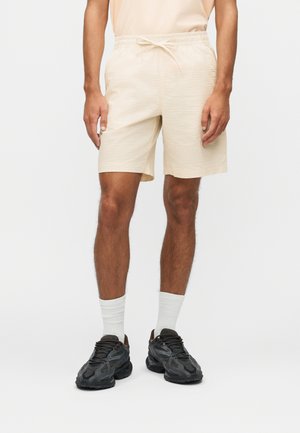 Moški v bež teksturiranih kratkih hlačah z elastičnim pasom, belimi nogavicami in črnimi športnimi supergami stoji pred enobarvnim ozadjem.