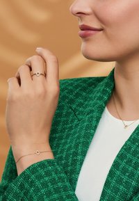 Kultaketju, jossa on soikea muotoilu ja pieni jalokivi, yhdistettynä kultaketjubaalaan ja herkän kaulaketjun kanssa, pukeutuneena vihreään tekstuuroituun bleiseriin.