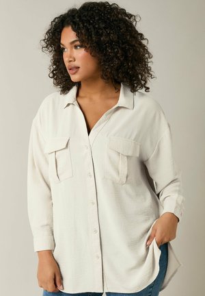 Camisa de botones de color beige claro con dos bolsillos en el pecho, corte holgado y mangas largas, que presenta un tejido texturizado y un dobladillo curvado.