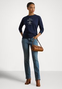 Μπλε σκούρο φούτερ με κεντημένο λογότυπο, μανίκια με μανσέτες, συνδυασμένο με τζιν ανοιχτού χρώματος και καφέ τσάντα. Το μοντέλο φοράει καφέ μπότες με τακούνι.