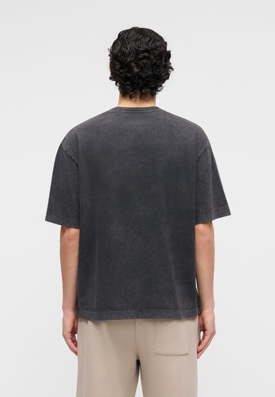 Άτομο με σγουρά σκούρα μαλλιά, ντυμένο με χαλαρό, ξεθωριασμένο μαύρο κοντομάνικο πουκάμισο και μπεζ παντελόνι, γυρισμένο πλάτη σε απλό φωτεινό φόντο.