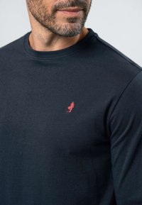 Homme portant un t-shirt bleu marine foncé à col rond avec un petit logo de plongeur rouge brodé sur la poitrine gauche, montrant une partie du visage et une barbe.