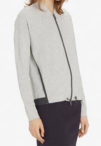 Grijze ritsjack met lange mouwen, gemaakt van zacht materiaal, met een zwarte zoom en trekkoorddetail bij de taille. Gladde textuur.