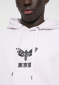 Ροζ φούτερ με λευκές κορδόνες, διαθέτει γραφικό με μια μαύρη στάθμη και ιαπωνικό κείμενο τυπωμένο μπροστά. Υλικό από μείγμα βαμβακιού.