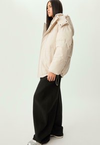 Kvinna som vänder åt vänster bär en beige puffjacka, vida svarta byxor och silverfärgade sneakers mot en enfärgad ljus bakgrund.