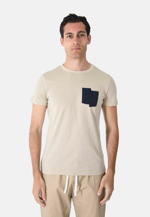 Uomo che indossa una maglietta beige a maniche corte con tasca rettangolare blu scuro e pantaloni beige con coulisse, in piedi su sfondo bianco.