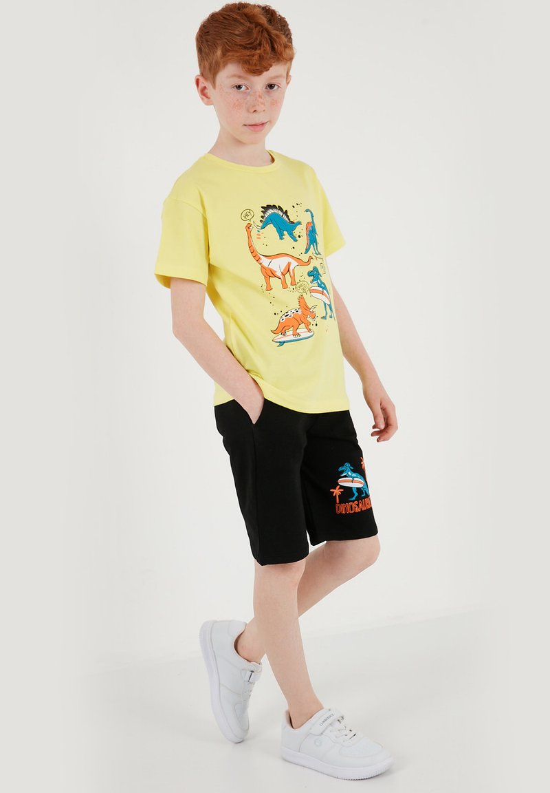 Garçon aux cheveux roux portant un t-shirt jaune avec un imprimé de dinosaure et un short noir avec un motif de dinosaure, posant les mains dans les poches et des baskets blanches.