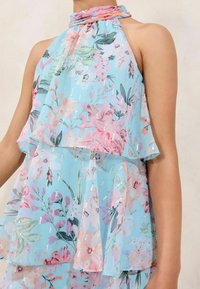 Hellbluse mit mehreren Lagen und floralem Druck in Pink und Grün, mit Neckholder und zarten, schimmernden Verzierungen überall.