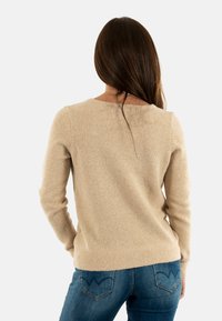 Maglione lavorato a maglia beige con collo rotondo, maniche lunghe e orlo a coste. Abbinato a jeans blu con un design a forma di cuore per la tasca posteriore.