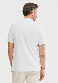 Biała koszulka polo z krótkim rękawem o gładkiej fakturze, z klasycznym kołnierzykiem i prostym dołem. Widoczne najważniejsze tatuaże na ramieniu.