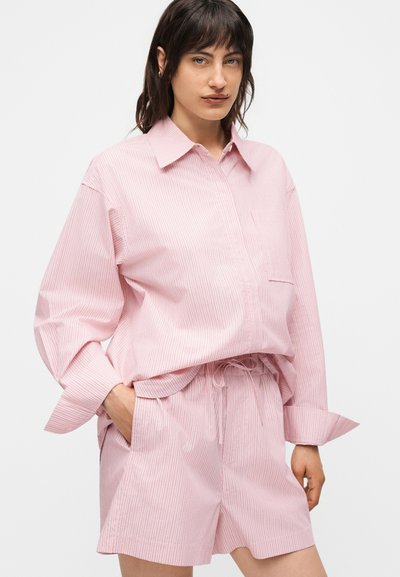 Γυναίκα που φορά χαλαρό ροζ πουκάμισο και ταιριαστό σορτς με λεπτές κάθετες ρίγες, χέρια στις τσέπες, ουδέτερο φόντο.