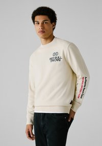 Crème sweatshirt met "RED BULL RACING" graphic, geribbelde kraag en manchetten, met een rode tekstaccent op de linkermouw. Draagt bij donkere broek.