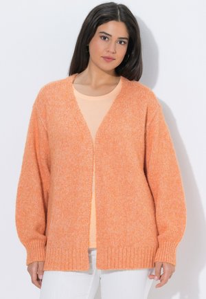 Giovane donna con capelli lunghi e scuri che indossa un cardigan arancione morbido e ampio sopra una maglietta pesca e pantaloni bianchi, su uno sfondo neutro.