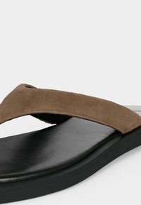 Bride en daim marron avec semelle intérieure rembourrée noire et semelle en caoutchouc noire sur une sandale minimaliste à bout ouvert.