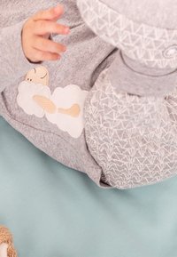 Bébé portant des vêtements gris avec une broderie de nuage et de personnage de dessin animé, allongé sur une surface bleue, une main levée et les pieds visibles.