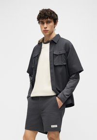 Νεαρός άνδρας με σγουρά μαλλιά φοράει λευκά και γκρι στρωμένα τοπ, σκούρα γκρι σορτς με λογότυπο Columbia, στέκεται με τα χέρια στις τσέπες.