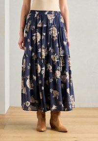 Marineblå blomster-maxikjole med lagdelt design, elastisk liv, og lett stoff; accentuert med beige og rustfargede blomster, kombinert med brune ankelstøvler.