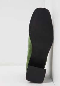 Vue de la semelle d'une chaussure en daim verte avec une semelle en caoutchouc lisse noire et un talon carré sur fond blanc.